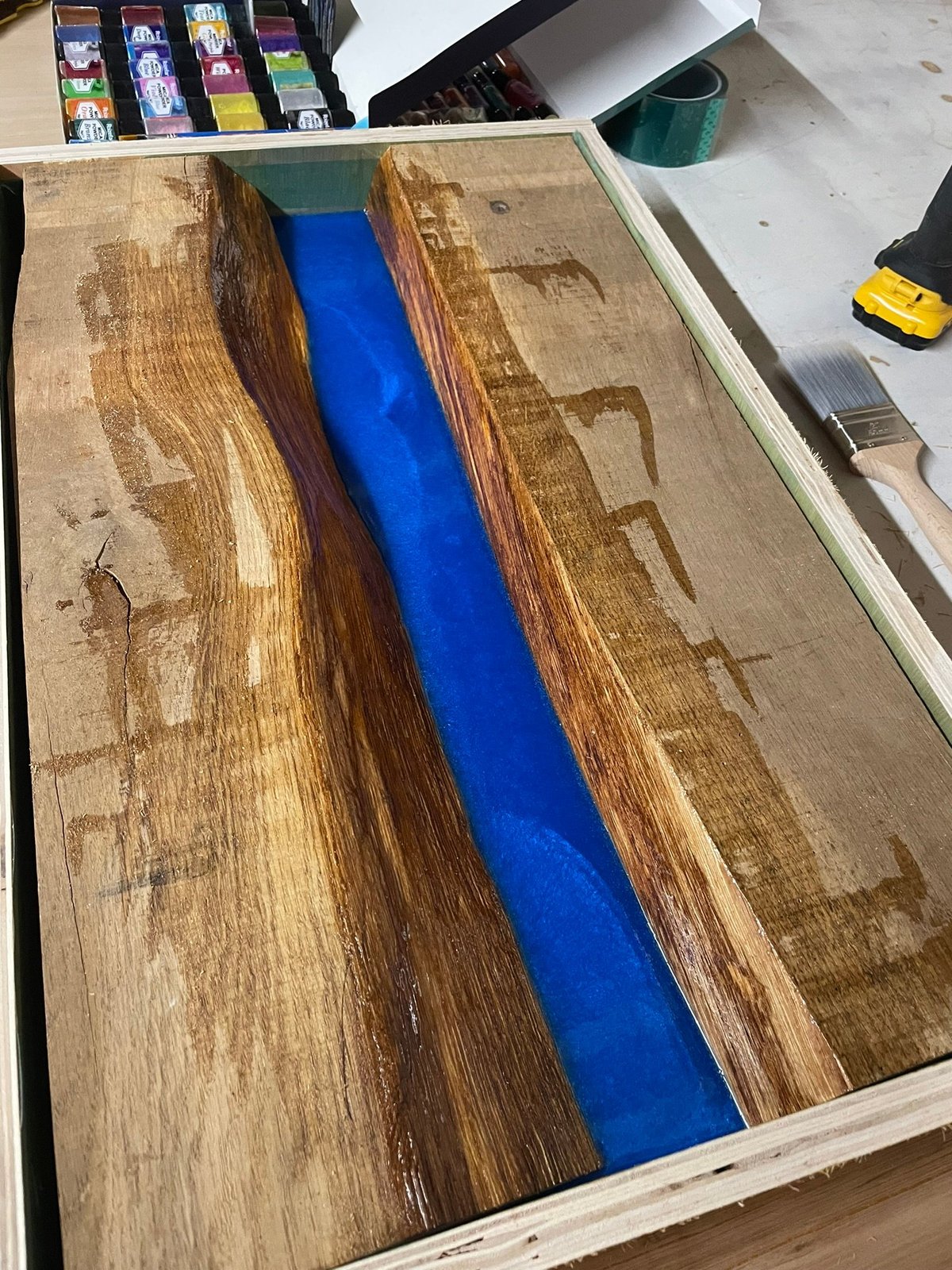 Wooden board with deep blue epoxy resin river running down center, surrounded by art supplies and tools on workbench