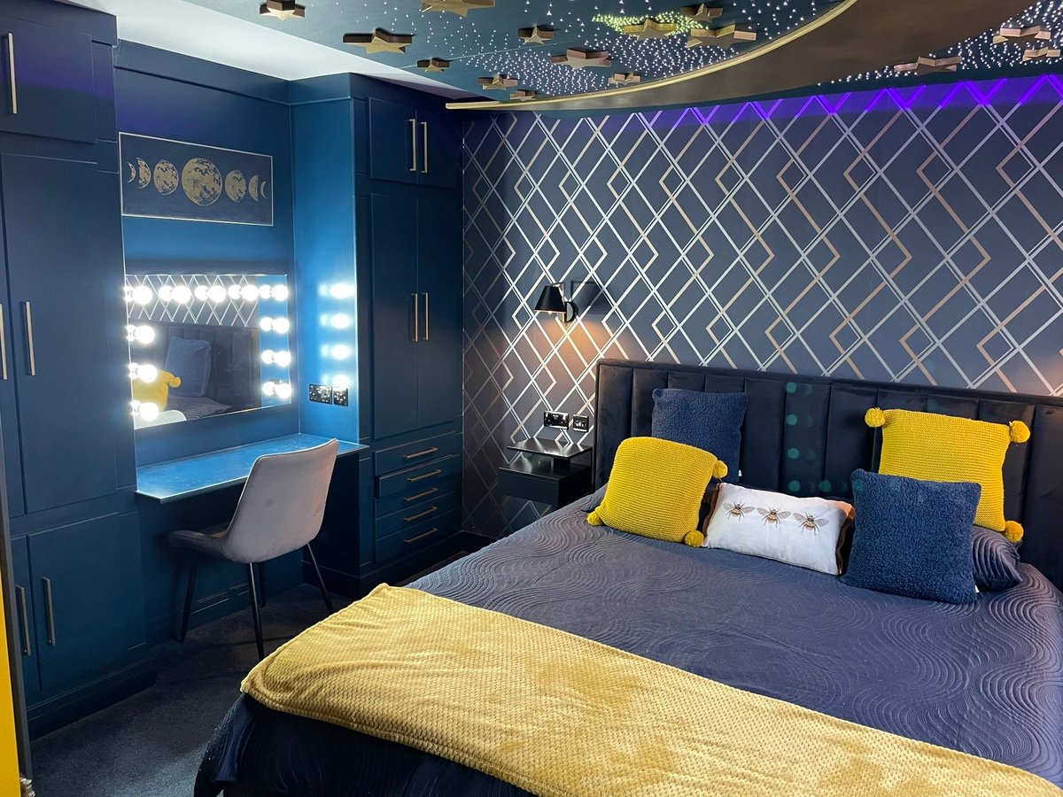 Modern bedroom with navy blue walls, built-in desk with LED lights, yellow and navy accents, patterned headboard wall, and geometric ceiling detail